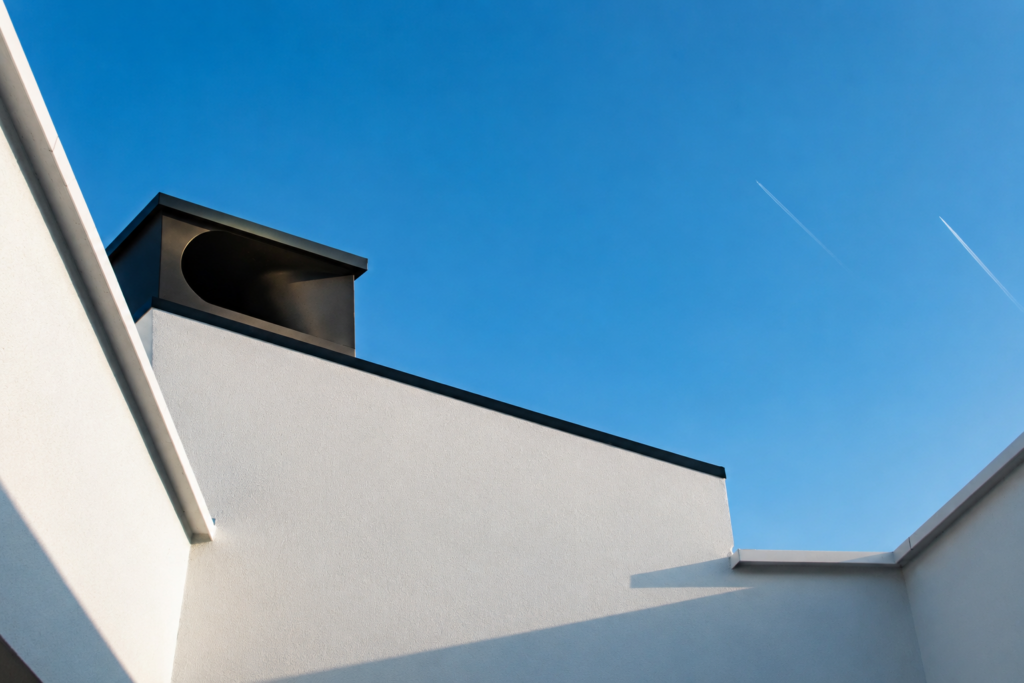 Détail architectural contemporain réalisé par Loyd Architects à Kain dans le Hainaut, mettant en valeur une façade minimaliste blanche et une cheminée design noire sous un ciel bleu lumineux.